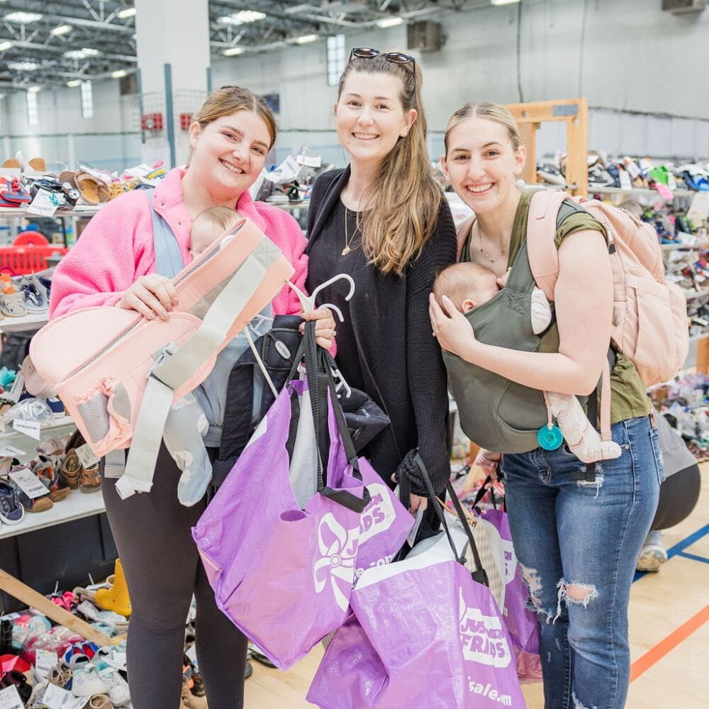 A mom with a large JBF shopping bag on her shoulder stands beside her husband who wears their toddler at a JBF sale.